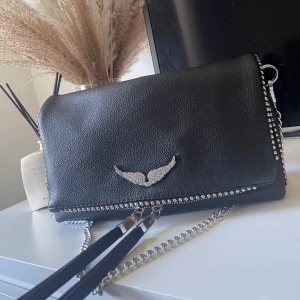 zadig Voltaire väska - Säljer min Zadig Voltaire väska (ej äkta) Väskan är köpt utomlands för runt 400kr och är i ett bra skick med endast missfärgning på dragkedjorna framtill på väskan och även på dom runda silver detaljerna som sitter på kanten på fram facket (se bild tre och fyra)  Ska ha ett till axelband till väskan som man kan byta emellan, skickar med de om jag hittar på de🌸