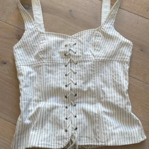 Vit randig korsett med snörning från zara - Supersnygg vit korsett-topp med tunna svarta ränder och breda axelband. Framsidan har cool snörningsdetalj och baksidan har dragkedja för enkel påklädning. Figurnära passform och perfekt för att lyfta vilken outfit som helst.