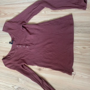 Ribbad långärmad tröja - Säljer en ribbad långärmad tröja från Pieces i en snygg vinröd nyans. Tröjan har rund halsringning och dekorativa knappar framtill. Perfekt för lager-på-lager och chill vardagsstil.