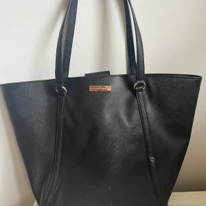 Svart axelväska från Tommy Hilfiger - Stilren svart axelväska från Tommy Hilfiger med guldiga metalldetaljer och logoplatta framtill. Väskan är rymlig med innerfack och dragkedja, perfekt för att bära allt du behöver. Tillverkad i strukturerat skinnliknande material och har dubbla axelband. Liten vit fläck på sidan som är lätt att gnugga bort med rätta medel.