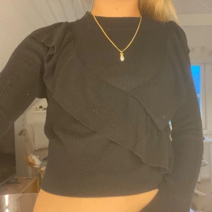 Svart stickad tröja med volang  - Supersnygg svart stickad tröja från Gina Tricot i storlek XS. Tröjan har långa ärmar, ribbad struktur och cool volangdetalj som går diagonalt över framsidan. Perfekt till höst och vinter för en trendig look.