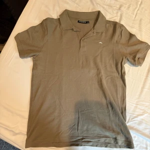 Grön pikétröja från J.Lindeberg - Snygg beige pikétröja från J.Lindeberg med klassisk krage och korta ärmar. Tröjan har en liten vit logga broderad på bröstet och knäppning framtill. Perfekt för en clean och stilren look.