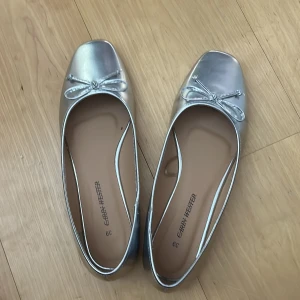 Silvriga ballerinaskor med rosett - Snygga ballerinaskor i metallic-silver. Jag köpte dem här på Plick men den var tyvärr för stora. Skorna har fyrkantig tå, platt sula och en söt liten rosett framtill. Perfekta för dig som vill sticka ut med en glansig touch. Tillverkade i syntetmaterial.