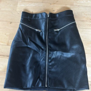 Svart skinnkjol med dragkedjor - Snygg svart kort skinnkjol från H&M  med coola silverfärgade dragkedjor framtill. Kjolen har en rak passform och markerad midja. Storlek 32 och i fint skick. Dock en liten inbuktning i skinet se bild 4. Den är väldigt liten och syns knappt. KAN ABSOLUT GÅ NER I PRIS VID SNABB AFFÄR ELLER BUNDLE!