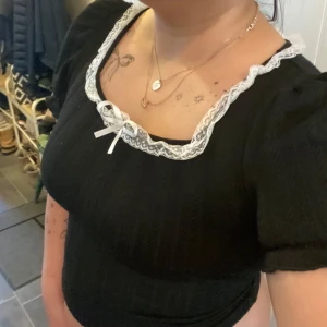 Svart topp med spets och rosett - Supersöt svart topp med korta puffärmar, vit spetskant runt fyrkantig ringning och en liten rosett framtill. Stickad känsla i materialet och figurnära passform. Klippte tyvärr bort lappen så minns inte vilken storlek men köpte det på yesstyle. Den passar storlek s eller m. Om du har storlek m så sitter den figurnära 