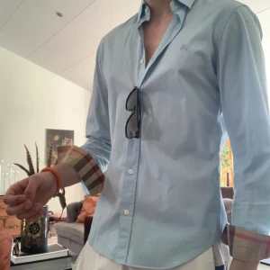 Ljusblå skjorta från Burberry Brit XS - Snygg ljusblå skjorta från Burberry Brit i klassisk modell med knappar hela vägen och button down-krage. Ikoniskt rutigt tyg på insidan av manschetterna. Tillverkad i bomull och passar perfekt till en stilren look.