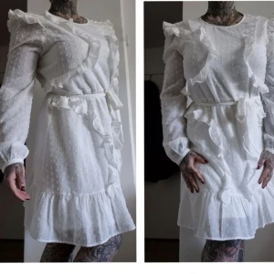 Vit volangklänning med bälte - Superfin vit kortklänning med volanger och knytbälte i midjan. Klänningen har långärmade ärmar, strukturmönster och volangdetaljer längs axlar och kjol. Perfekt för dig som gillar romantisk och söt stil.