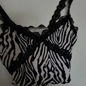 Zebramönstrad spetslinnetopp - Säljer en snygg zebramönstrad linnetopp med svarta spetsdetaljer längs urringning och axelband. Toppen är vit och svart, har smala axelband och är perfekt för sommaren! Materialet känns mjukt och följsamt.🤍