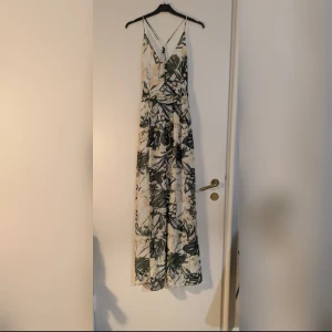 Långklänning med dekorativa knappar - Superfin långklänning med mönster i grönt, beige och vitt. Klänningen har smala axelband, v-ringning och dekorativa knappar framtill. Snyggt flow i kjolen och öppen rygg med tunna band.