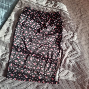 Svart blommig kjol - Svart kjol som går unger till knät med små rosa och vita blommor över hela tyget. Kjolen har en snygg slits på sidan som ger en cool touch. Perfekt för dig som gillar romantiska mönster och vill ha något som sticker ut lite extra. Kjolen mäter ca 69 cm från topp till botten.