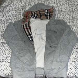 Grå Burberry hoodie med rutig huva - Säljer en grå hoodie från Burberry med dragkedja och klassiskt rutigt foder i huvan. Tröjan har snörning vid huvan, långa ärmar och ribbade muddar. Perfekt för dig som gillar stilrena och exklusiva streetwear-plagg.