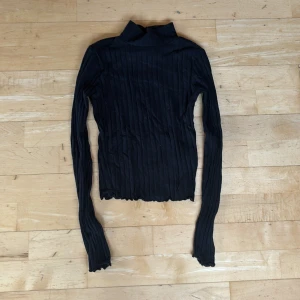Svart ribbad polotröja Gina Tricot XS - Svart ribbad polotröja från Gina Tricot i storlek XS. Tröjan har lång ärm  och hög hals. Materialet är stretchigt och mjukt, perfekt för lager-på-lager. Fina vågiga kanter vid ärmslut och nederkant ger en extra detalj. Lite vita katthår på som såklart tas bort innnan den postas. KAN ABSOLUT GÅ NER I PRIS VID SNABB AFFÄR ELLER BUNDLE!