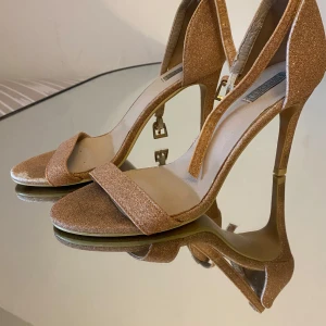Glittriga guldiga pumps med klack - Snygga pumps i glittrigt guld med öppen tå och hög klack. Skorna har en elegant rem över tårna och smal ankelrem med spänne. Perfekta för att lyfta din outfit med extra glamour och stil.