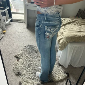 Lågmidjade jeans - Säljer ett par ljusblå jeans från Telepatía med snygg bootcut passform. Jeansen har låga fickor bak med broderade detaljer, paljetter och kontrastsömmar. Klassisk femficksmodell och låg midja. Perfekta för dig som vill sticka ut lite extra.