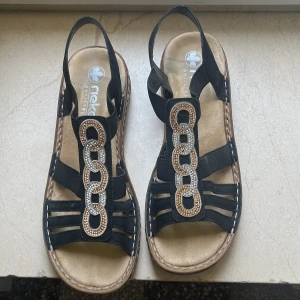 Svarta sandaler från Rieker med kedjedetalj - Snygga svarta sandaler från Rieker med breda remmar och dekorativa kedjelänkar med glittriga stenar framtill. Mjuk innersula och brun sula med kontrastsöm. Perfekta för varma dagar när du vill ha både stil och komfort.