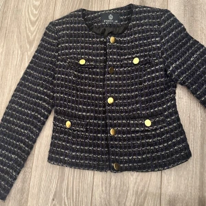 Mörkblå kavaj från Stockh LM, stl 34 - Snygg mörkblå kavaj från Stockholm  i klassiskt rutigt mönster. Jackan har guldiga knappar framtill och på fickorna, rund halsringning och lång ärm. Materialet är strukturerat och känns lyxigt, perfekt för att lyfta din outfit. Använd 1 gång