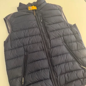 Navy dunväst från Parajumpers - Snygg svart dunväst från Parajumpers med quiltad design och gul detalj vid kragen. Västen har två dragkedjefickor framtill och en vid bröstet  och hög krage. Insidan är grå och har märkets logga. Perfekt för lager-på-lager och chill stil. Köpte den till lillebrorsan där han bara använt den 3-4 gånger. Nypris 4499kr