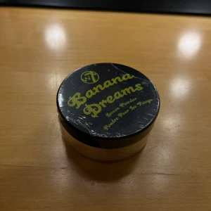 W7 Banana Dreams Loose Powder - Banana Dreams Loose Powder från W7 är ett löspuder i en ljusgul nyans som ger en matt och jämn finish. Perfekt för att fixera foundation och dämpa glans. Kommer i en rund plastburk med svart lock och gul text. Oöppnad