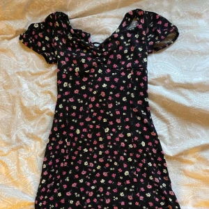 Svart blommig kortklänning med snörning - Supersöt svart kortklänning med små rosa och gula blommor. Klänningen har korta puffärmar, v-ringning och snygg snörning både fram och bak som ger en öppen rygg. Perfekt för sommarens alla tillfällen!