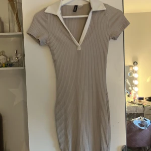 Beige ribbad kortklänning H&M Divided - Supersnygg ribbad kortklänning från H&M Divided i beige med vit krage och v-ringning. Klänningen har korta ärmar och figurnära passform, perfekt för en trendig och sportig look. Materialet är stretchigt och bekvämt.