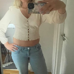 Beige croppad blus med knappar - Superfin beige croppad blus med v-ringning och långa puffärmar. Smockad midja och ärmslut ger en snygg passform. Snygga knappar framtill och lätt volangkant nedtill. Perfekt till jeans eller kjol för en trendig look.