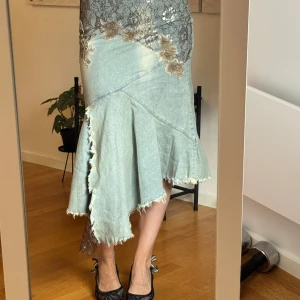 Asymmetrisk jeanskjol med spetsdetaljer - Unik ljusblå långkjol i jeans med asymmetrisk volangkant och råa kanter. Kjolen har svarta och beige spetsapplikationer med blommor upptill och en cool slits med spetsinslag. Perfekt för dig som vill sticka ut med en edgy och kreativ stil.