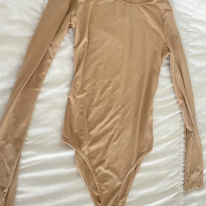 Två långärmade bodys i beige och brun - Säljer två stilrena långärmade bodys i slät, stretchig syntet. Ena är beige och den andra är brun, båda med rund hals och figurnära passform. Perfekta att styla med jeans eller kjol för en clean look. 