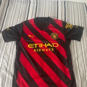 Manchester City tröja  - Snygg Manchester City bortatröja med Haaland 9 på ryggen. Svart och röd med gula detaljer, diagonalrandigt mönster och klubbmärke på bröstet. Puma-logga och Etihad Airways tryck framtill. Perfekt för dig som älskar fotboll och vill sticka ut.