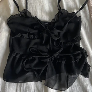 Svart volangtop från PrettyLittleThing - Supersöt svart linnetopp från PrettyLittleThing med tunna axelband, volanger och flera knytband framtill.  Passade inte mig💕