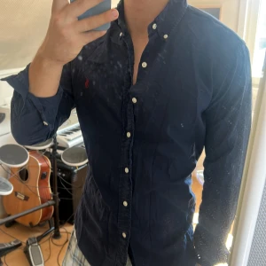 Mörkblå skjorta från Ralph Lauren - Säljer en mörkblå skjorta från Ralph Lauren med klassisk röd broderad logga på bröstet. Skjortan har lång ärm, knappar hela vägen och button-down krage. Tillverkad i mjuk bomull och har en normal passform. Perfekt för dig som gillar stilrena och tidlösa plagg.