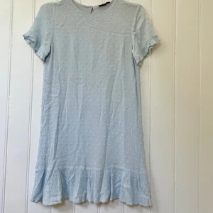 Ljusblå kortklänning från LMTD - Supersöt ljusblå kortklänning från LMTD med diskret prickigt strukturmönster, volangkant nertill och korta ärmar med liten volang. Perfekt för sommarens alla tillfällen. Nyckelhålsöppning med knapp i nacken. Lätt och luftig känsla.