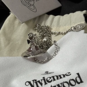 Vivienne Westwood halsband med rosa sten - Ikoniskt halsband från Vivienne Westwood i silverfärg med ett hängsmycke i form av deras klassiska orb. Hängsmycket har en stor ljusrosa sten i mitten och detaljer runt om. Kedjan är tunn och har märkets logga ingraverad på brickan vid låset.