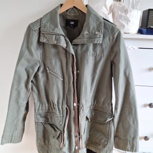 Grön parkajacka från H&M, strl 38 - Snygg grön parkajacka från H&M i storlek 38. Jackan har flera fickor med lock och dragkedja, justerbar midja med snörning och coola detaljer vid axlarna. Perfekt för vår och höst, tillverkad i bomull för en skön känsla. En av fickorna har lossnat i sömmarna men den händiga kan lätt sy fast den igen!