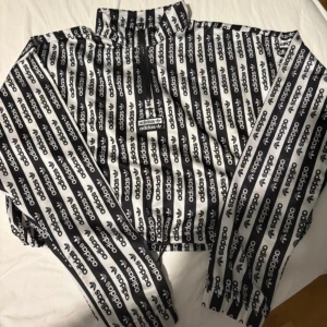 Svartvit vindjacka från Adidas - Säljer en svartvit vindjacka från Adidas med heltryck av loggan i ränder över hela jackan. Jackan har hög krage, dragkedja framtill och elastiska muddar. Perfekt för dig som vill sticka ut med en sportig och trendig look.