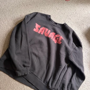 Svart sweatshirt  - Cool svart sweatshirt med stort rött SAVAGE-tryck på bröstet. Tröjan har rund hals, långa ärmar och ribbade muddar vid ärmslut. Finns en liten vit fläck men som knappt syns
