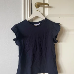 Mörkblå volangtopp från Gina Tricot - Supersöt mörkblå topp från Gina Tricot med volangdetaljer på axlarna. Modellen är ärmlös och har en klassisk rund halsringning. Tillverkad i mjuk bomull som känns skön mot huden. Perfekt för dig som gillar en enkel men ändå trendig look. Storlek 146/152.