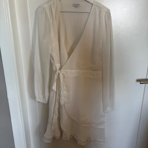 Vit omlottklänning från Bubbleroom - Säljer en elegant vit omlottklänning från Bubbleroom med långa ärmar och volangdetalj nertill. Klänningen har knyt i midjan och en v-ringad halsringning. Perfekt för sommarens tillfällen eller studentfirande!