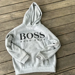 Grå hoodie från Hugo Boss - Säljer en grå hoodie från Hugo Boss med stor svart logga framtill. Tröjan har huva och en klassisk känguruficka. Perfekt för en avslappnad och stilren look. Pris kan diskuteras 
