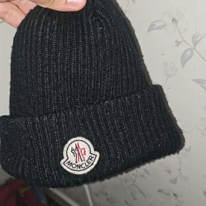 Svart ribbstickad mössa från Moncler - Svart ribbstickad mössa från Moncler med uppvikt kant och klassisk Moncler-logga framtill. Tillverkad i mjukt material som håller värmen. Perfekt accessoar för kyliga dagar.