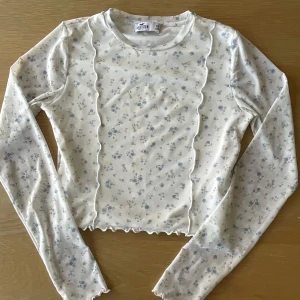 Vit blommig meshtröja från Hollister - Säljer en söt, croppad långärmad meshtröja från Hollister i vitt med små blå blommor. Tröjan har vågiga kanter och är figurnära. Perfekt att styla med jeans eller kjol!