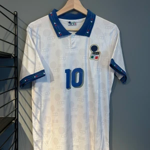 Italien fotbollströja nummer 10 R. Baggio - Klassisk vit fotbollströja från Italiens landslag med blå detaljer, nummer 10 och R. Baggio på ryggen. Blå krage och ärmslut med mönster, samt Italiens emblem på bröstet. Perfekt för fans av italiensk fotboll. En replika helt ny i S