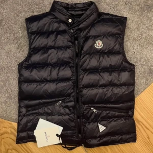 Svart dunväst från Moncler äkta - Säljer en svart, glansig dunväst från Moncler med ståkrage och dragkedja framtill. Västen har två snedställda fickor med dragkedja och Moncler-logga på bröstet. Perfekt för lager-på-lager och stilren look.