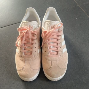 Adidas Gazelle i ljusrosa mocka - Säljer ett par Adidas Gazelle sneakers i ljusrosa mocka med vita detaljer och klassiska tre ränder på sidan. Skorna har snörning och vit sula, finns även vita snören som skickas med. Använda några gånger då jag tyvärr växte ur den snabbt, men den ljusrosa färgen är kvar. Lite missfärgade längst fram. Storlek 36 och 1/3❤️❤️ skriv vid frågor❤️❤️