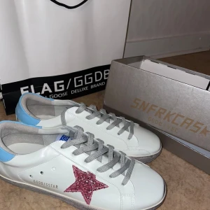 Golden Goose Sneakers – Super-Star, Vita med Rosa stjärn - Säljer ett par Golden Goose Deluxe Brand Super-Star sneakers. Klassiska vita lädersneakers med glitterrosa  stjärna på sidan, grå skosnören och blå häl. Skorna är i mycket gott skick och kommer i originalkartong och påse 