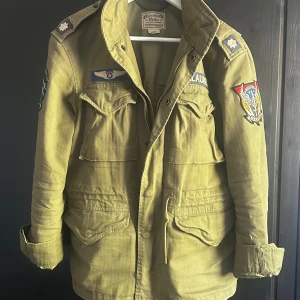 Fieldjacket från Polo Ralph Lauren - Säljer en olivgrön militärjacka från Polo Ralph Lauren med flera coola patchar på ärmar och bröst. Jackan har dragkedja, stora fickor framtill och klassisk krage. Perfekt för dig som gillar utility-stil och streetwear.