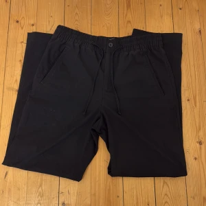 Svarta byxor med resårmidja från H&M - Svarta byxor från H&M med elastisk midja och dragsko. Byxorna har raka ben, sidofickor och en bakficka. Perfekta för en avslappnad stil och enkel att matcha med olika plagg.