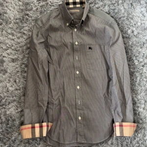 Randig skjorta från Burberry Brit - Snygg grå och vit randig skjorta från Burberry Brit med klassiska rutiga detaljer på insidan av manschetterna. Skjortan har knappar framtill, bröstficka och lång ärm. Tillverkad i 100% bomull.