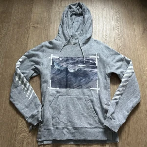 Grå hoodie från Off-White | ev byte - Säljer en grå hoodie från Off-White med tryck av vågor på bröstet och vita diagonala ränder på ärmar och rygg. Tröjan har huva och magficka, perfekt för en avslappnad streetwear-look.