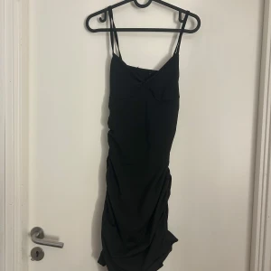 Svart tight kortklänning med smala axelband - Säljer en svart tight kortklänning med smala axelband och draperad detalj på sidan. Klänningen har en enkel och stilren design som passar perfekt till sommarens alla tillfällen.