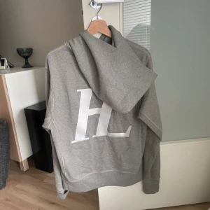 Helmut Lang Hoodie M - Säljer en stilren grå hoodie från Helmut Lant  med stor vit HL-logga på ryggen och liten logga fram. Tröjan har huva och magficka, perfekt för en avslappnad look.. sitter väldigt bra och det är väldigt fet